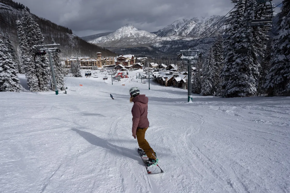 An energetic action shot of a snowboarder carving down a snowy slope with the mountain range in the background, ski resort buildings, and ski lifts visible below. The image radiates excitement and the thrill of winter sports amidst breathtaking alpine scenery.