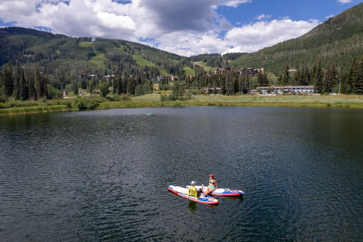 Two people paddleboard across a tranquil lake with forested mountains in the background. The scene is playful and serene, suggesting nearby outdoor activities and shared experiences in nature. The golden hour lighting enhances the sense of romance or adventure.
