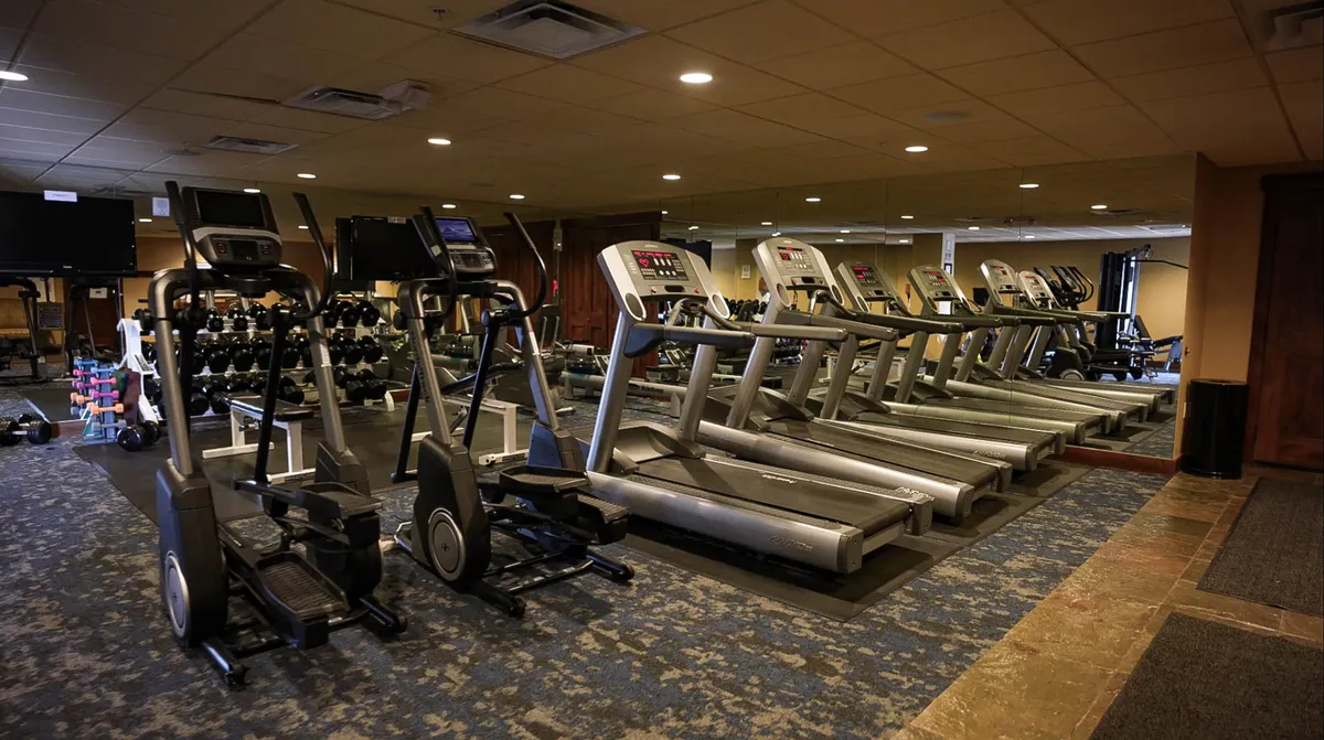 An inviting interior fitness center equipped with treadmills, ellipticals, a weight lifting area, and mirrored walls. The scene suggests guests actively working out, participating in group classes, or achieving their fitness goals in a bright, motivational environment.