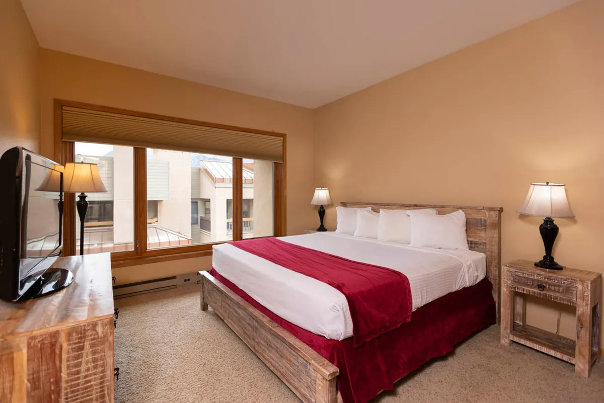 A spacious bedroom featuring a plush king-sized bed with crisp linens, wooden bedside tables, a television, and large windows that flood the room with natural light. The scene evokes a restful, inviting atmosphere ideal for relaxation and unwinding.