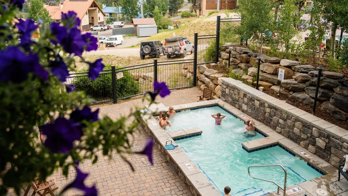 This exterior image features a luxurious hot tub and swimming pool, surrounded by natural beauty and vibrant flowers in the foreground. The scene captures guests relaxing in the hot tub, socializing by the pool, and enjoying the tranquility of the setting, with sunset hues illuminating the space.