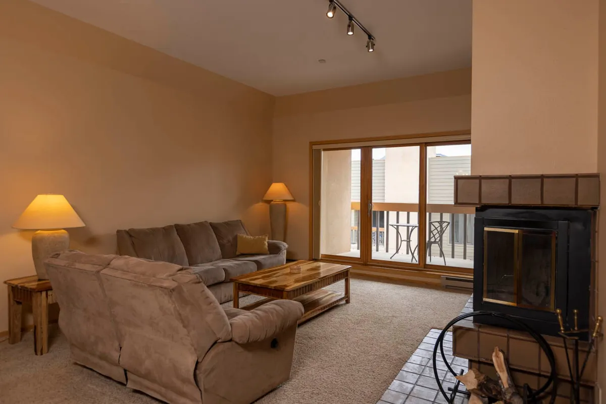 This image showcases a plush sofa arranged around a cozy wood fireplace, with a coffee table and floor lamp adding to the welcoming ambiance. A large window offers glimpses of the outdoors, suggesting mountain views and natural light that fills the space. The scene suggests relaxation, socializing, and comfort, perfectly inviting for après-ski gatherings.