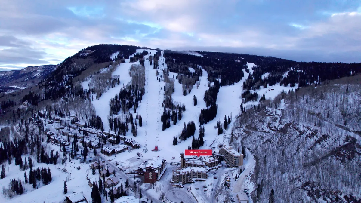 A sweeping exterior view of the ski slopes blanketed in snow with the Village Center nestled at the base, surrounded by majestic mountains and snow-covered trees. The scene captures the vibrant heart of the resort, with skiers and snowboarders enjoying the first runs of the day and warm lights emanating from the main plaza.