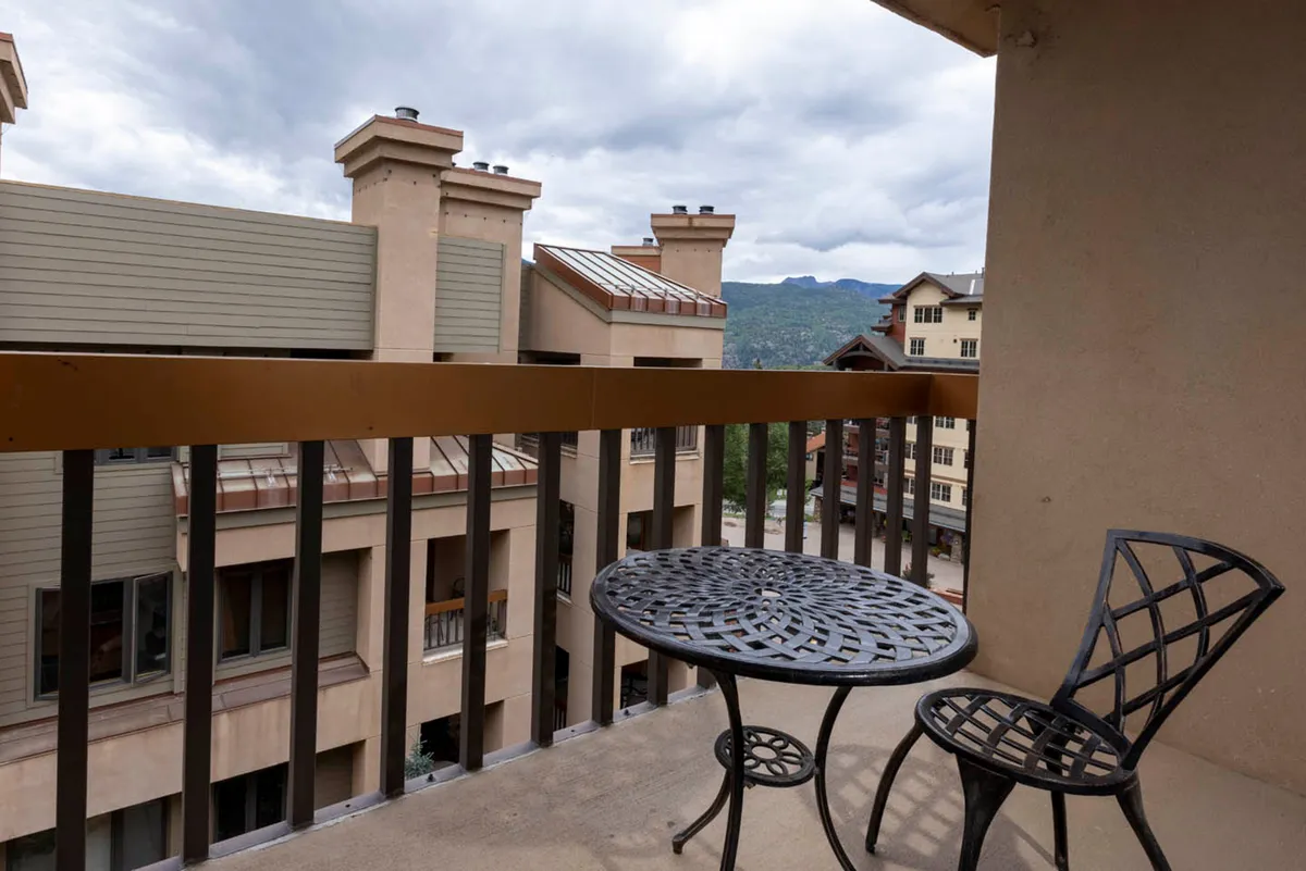 This exterior deck scene features a small round table and two chairs looking out over neighboring buildings and mountains. The image conveys a relaxed mood, perfect for enjoying a quiet breakfast, sipping coffee, or simply soaking in the views from the private deck.