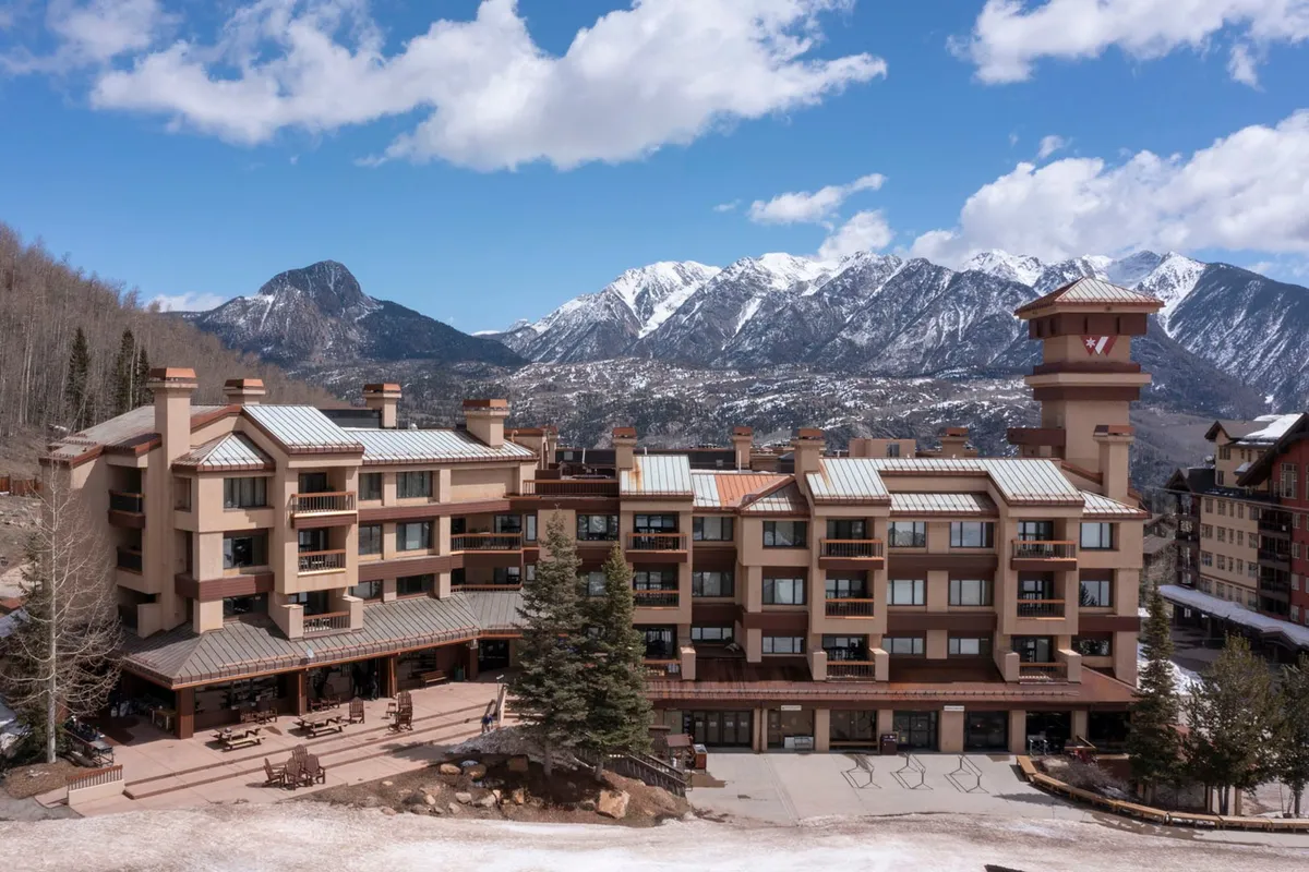 The image captures the main building of the property nestled against a stunning mountain backdrop. There’s an inviting outdoor seating area, perfect for gatherings, and the terrace offers sweeping views at sunset. The scene conveys an atmosphere of relaxation, adventure, and sociable energy, ideal for groups or couples.