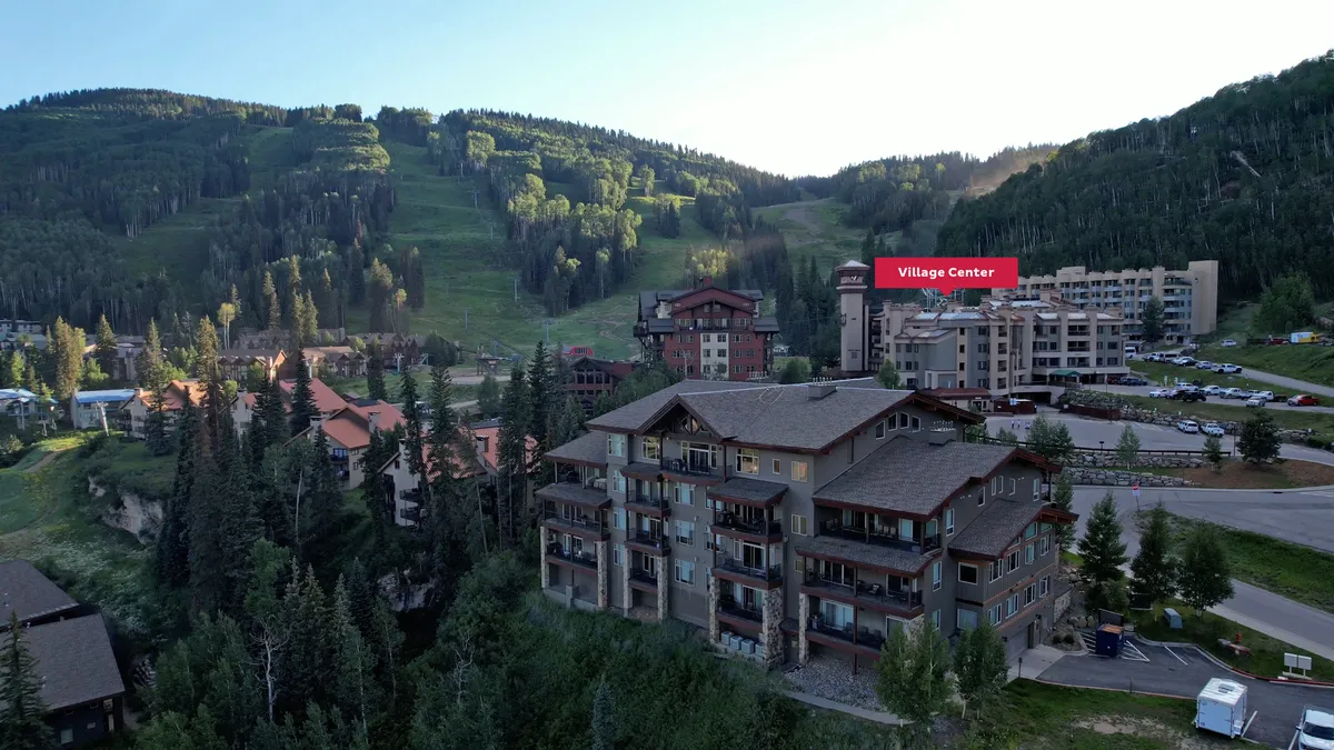 A wide exterior view featuring the Village Center, surrounding resort buildings, and parking area with rugged mountains in the distance. The scene highlights both the convenience of the location and the inviting, adventurous spirit of the village at sunset.