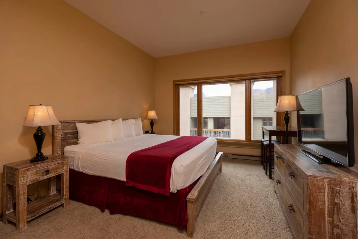 A serene bedroom arranged with another king-sized bed, nightstands with lamps, a television, and expansive windows offering mountain views. Evening light creates a peaceful, retreat-like setting.